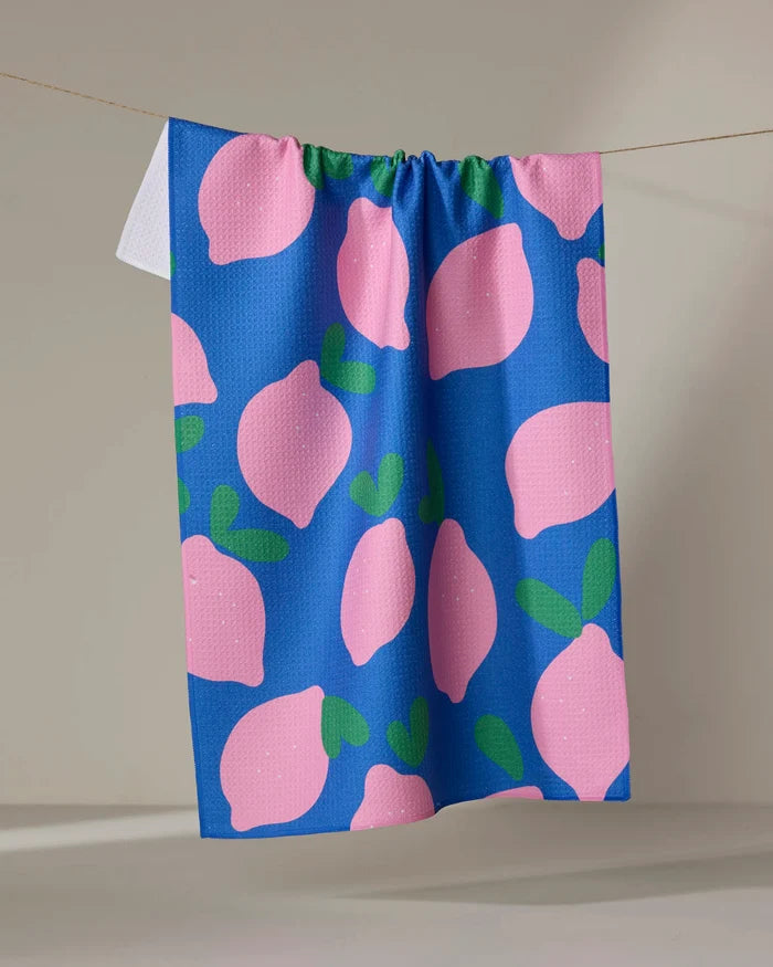 The Pink Lemons tea towel by Jennifer Bouron features large pink lemons with green leaves on a blue background and dries quickly.