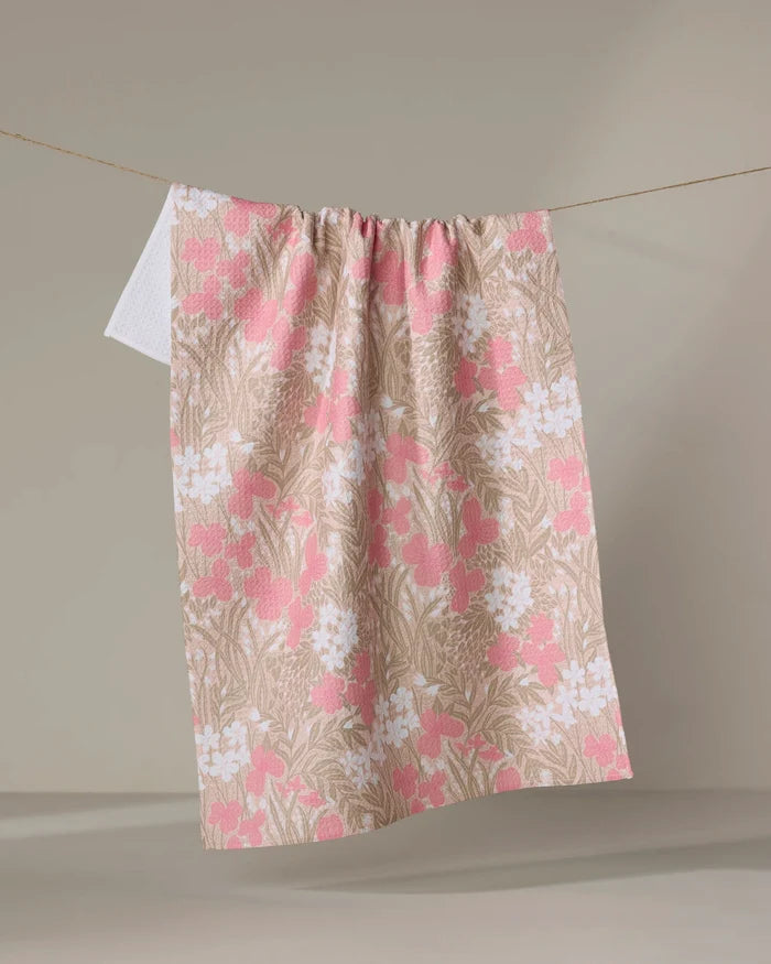 The "Pink Meadow" tea towel by Katie O'Shea, crafted from recycled materials, features a pink and white floral design and hangs from a clothesline against a light background.