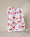 The GEOMETRY Pink Palms dish towel, featuring a vibrant pink and orange palm tree design, hangs on a line behind two clear ribbed glass cups set on a beige surface.