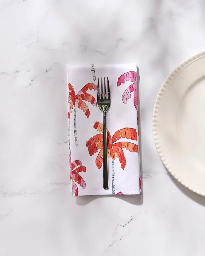 A silver fork rests on GEOMETRY’s Pink Palms eco-friendly napkin with pink and orange palm prints, placed on a white marble surface beside a white, scallop-edged plate from our Dinner Napkin Set.