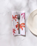 A silver fork rests on GEOMETRY’s Pink Palms eco-friendly napkin with pink and orange palm prints, placed on a white marble surface beside a white, scallop-edged plate from our Dinner Napkin Set.