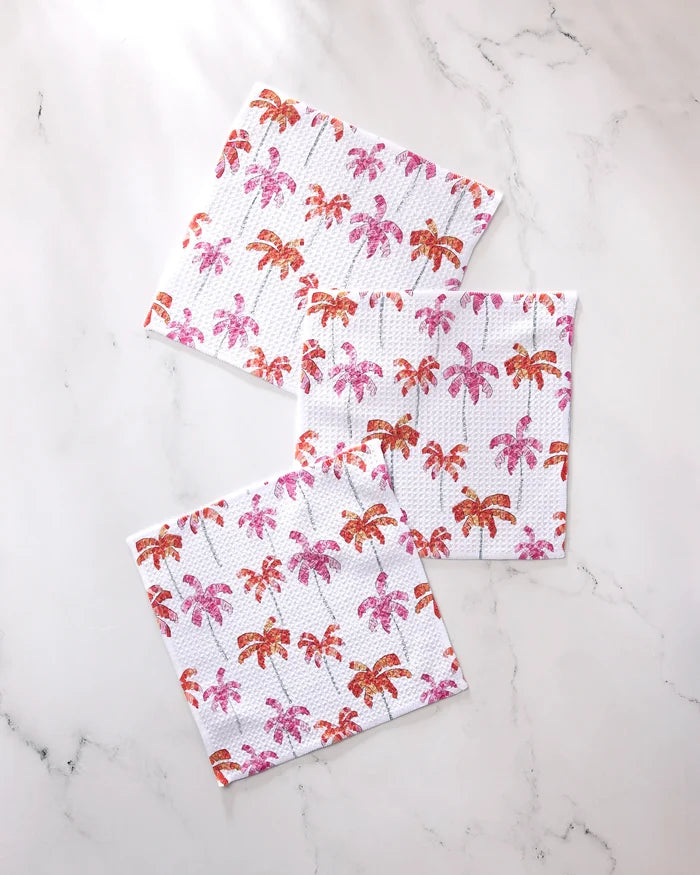 The GEOMETRY Pink Palms dishcloth set includes three super-absorbent towels, each adorned with pink and orange palm tree patterns on a white marble background.