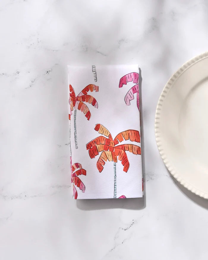 The GEOMETRY Pink Palms napkin, featuring pink and orange palm tree prints on white fabric, is folded neatly on a marble surface beside a scalloped plate—an eco-friendly choice for stylish table settings.