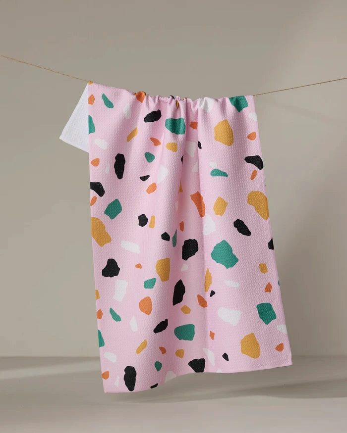 The Megan Roy Pink Terrazzo kitchen towel features a pink textured fabric with green, orange, yellow, white, and black terrazzo shapes. This eco-friendly accent brightens any kitchen with its playful, modern design.