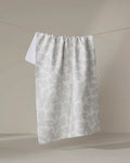 The Pixie Sage tea towel by Studio Jeannot, featuring a light blue-grey dotted pattern on white and crafted from recycled materials, hangs on a clothesline against a neutral beige background.