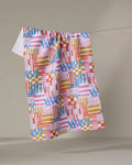 The "Playful Stripes" recycled kitchen towel by Claire Iglesias, featuring red, pink, blue, yellow, and white geometric squares and stripes, hangs on a string against a neutral background.