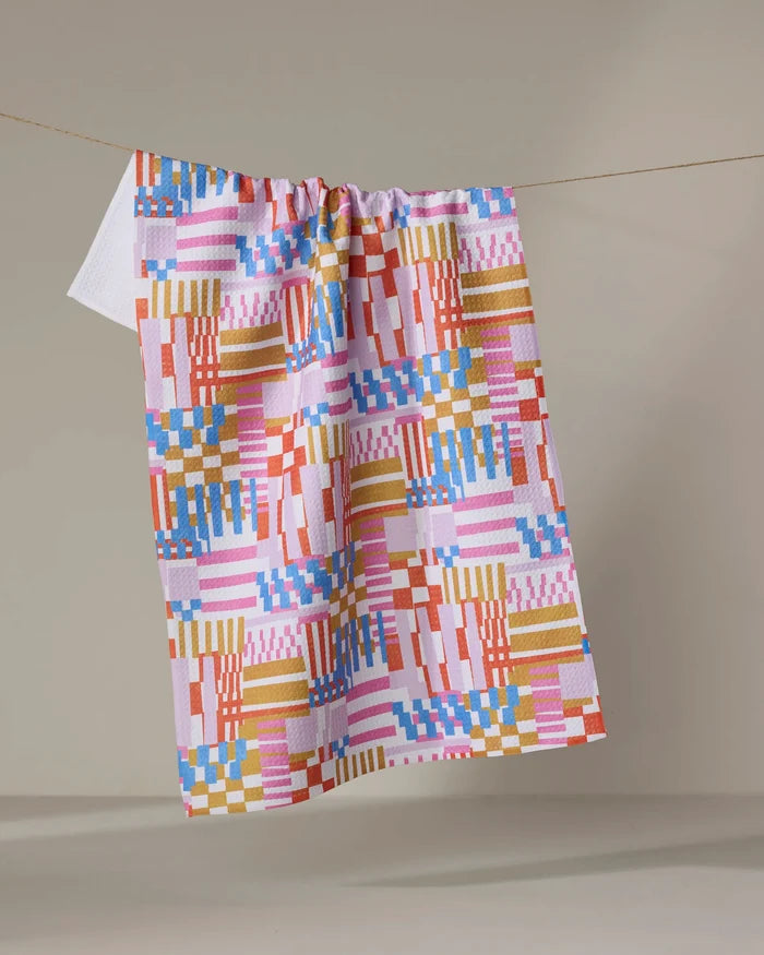 The "Playful Stripes" recycled kitchen towel by Claire Iglesias, featuring red, pink, blue, yellow, and white geometric squares and stripes, hangs on a string against a neutral background.