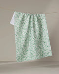 The Plumbago Bloom tea towel by Danelys Sidron, featuring a light green and white floral pattern, hangs on a clothesline against a plain beige background.