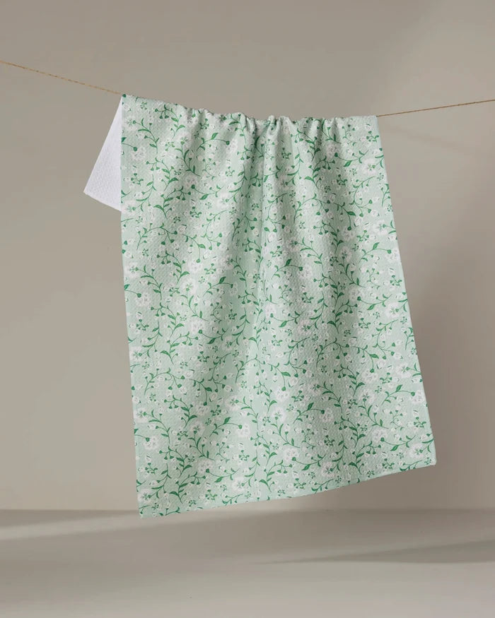 The Plumbago Bloom tea towel by Danelys Sidron, featuring a light green and white floral pattern, hangs on a clothesline against a plain beige background.