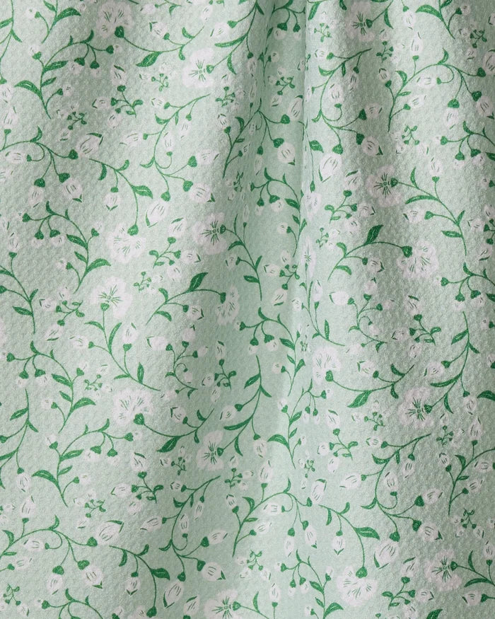 The Plumbago Bloom kitchen towel by Danelys Sidron features a light green, recycled fabric with a subtle texture and an elegant pattern of green vines and white flowers. Its gentle folds create light and shadow on the quick-drying surface.