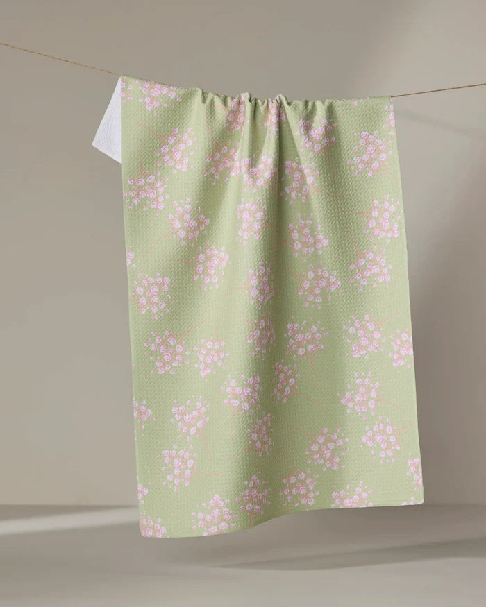 The Pocket of Posies kitchen towel by Sarah Reed, in light green with small pink and white floral patterns, hangs over a clothesline against a neutral background.