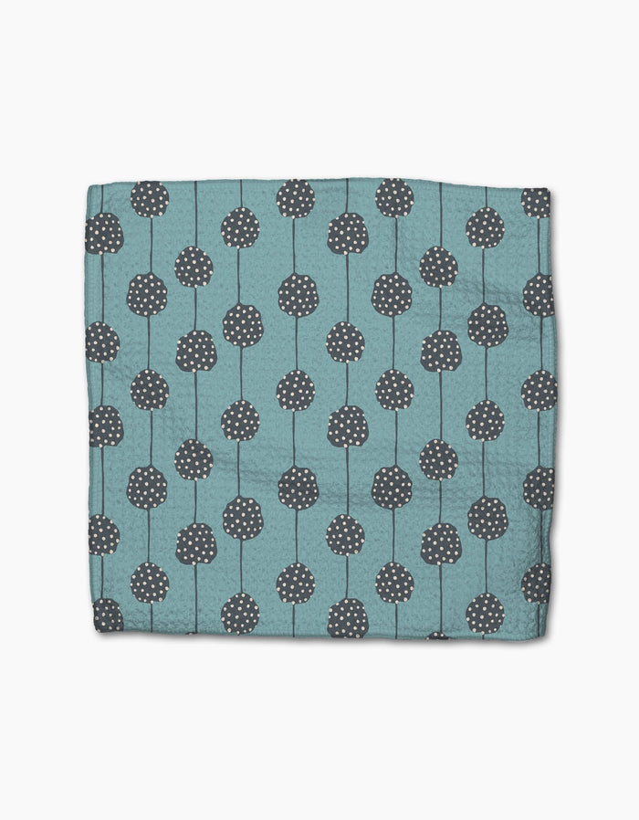 The Polka Dot Blooms napkin by Lizzie Clark is a folded teal cloth made from recycled materials, featuring dark gray circular patterns with white dots and vertical lines, displayed on a plain white background.