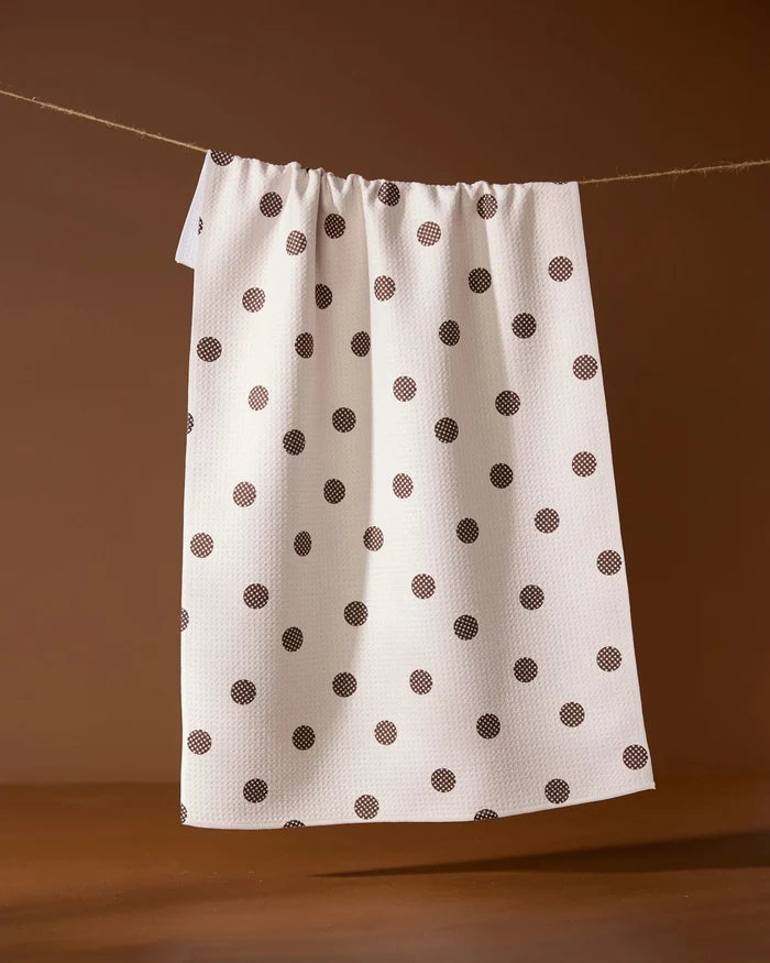 The Polka Picnic tea towel by Geometry, featuring black polka dots on white, hangs on a string against a brown background.