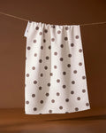 The Polka Picnic tea towel by Geometry, featuring black polka dots on white, hangs on a string against a brown background.