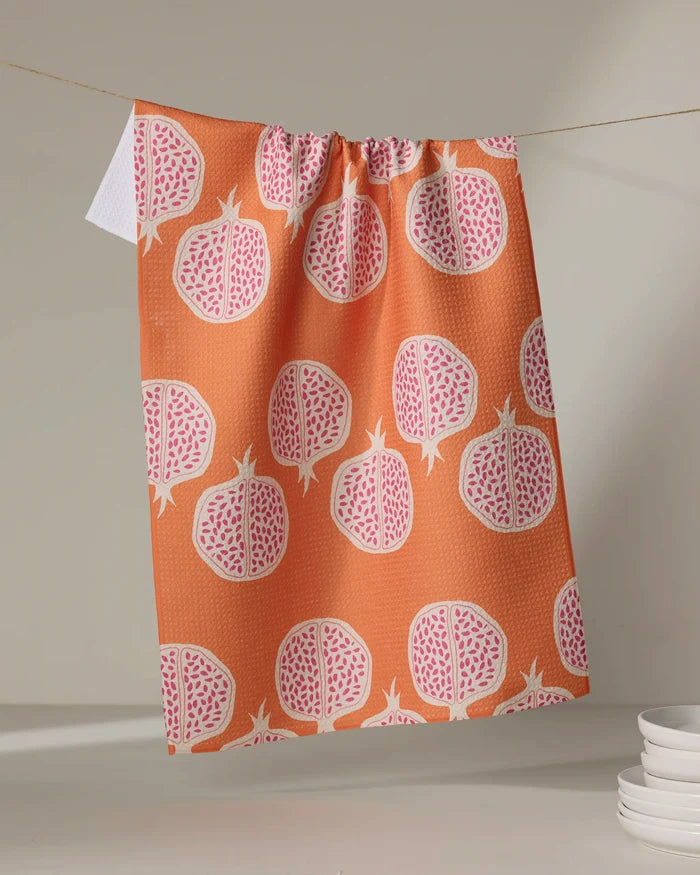 The Lizzie Clark Pomegranate Parade tea towel, made from quick-drying recycled materials and featuring a bright pomegranate pattern, hangs on a clothesline with its white tag visible. White plates are stacked in the lower right background.