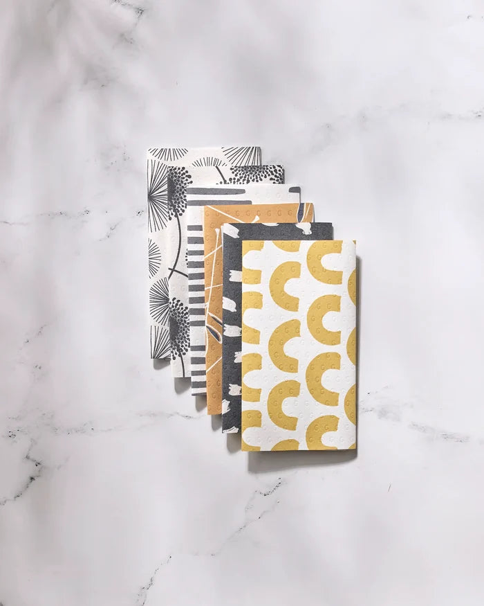 Four Pop Of Color reusable towels by GEOMETRY, featuring black, white, beige, and yellow geometric and abstract patterns, are folded in a staggered stack on white marble. Eco-friendly and stylish alternatives to paper towels.