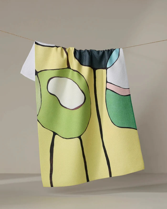 The "Poppies on Yellow" tea towel by Rachel Austin, featuring vibrant abstract shapes, hangs on a clothesline against a neutral backdrop.