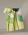 The "Poppies on Yellow" tea towel by Rachel Austin, featuring vibrant abstract shapes, hangs on a clothesline against a neutral backdrop.