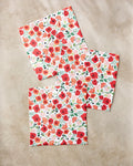 Three square pieces from Elisabeth Wing’s “Poppy Power” absorbent dishcloth set, featuring a red and orange floral pattern with green leaves and black accents, are arranged on a light beige surface.