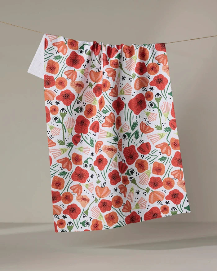 The "Poppy Power" quick-drying towel by Elisabeth Wing features vibrant red and orange poppies, green leaves, and abstract shapes, displayed on a clothesline against a neutral background.