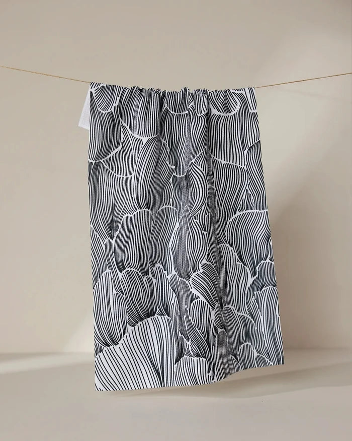 The Geometry Posh Petals Fresh Linen tea towel, featuring black and white abstract wavy lines, hangs on a string against a plain beige background.