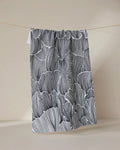 The Geometry Posh Petals Fresh Linen tea towel, featuring black and white abstract wavy lines, hangs on a string against a plain beige background.