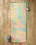 The "Positive Vibes" yoga mat by Sareka Unique, featuring a pastel abstract pattern, is laid on a wooden floor with a non-slip towel. Two gray blocks are at the bottom left and a white bottle sits at the top right.