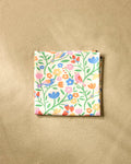 The Primrose Path beach blanket by Charlie Rowan Designs features a vibrant floral and bird pattern in pink, orange, blue, and yellow on quick-drying recycled fabric, shown folded on a neutral beige surface.