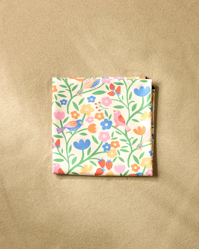 The Primrose Path beach blanket by Charlie Rowan Designs features a vibrant floral and bird pattern in pink, orange, blue, and yellow on quick-drying recycled fabric, shown folded on a neutral beige surface.