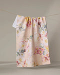 The Primroses tea towel by Pascale Dilger, featuring a colorful floral design in light pink and made from recycled materials, hangs on a clothesline against a neutral background.