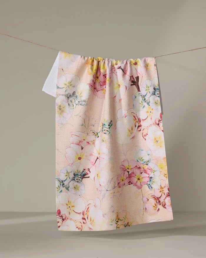 The Primroses tea towel by Pascale Dilger, featuring a colorful floral design in light pink and made from recycled materials, hangs on a clothesline against a neutral background.