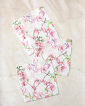 Three folded Pascale Dilger "Primroses" napkins, featuring a pink floral pattern with delicate branches and blossoms, are arranged on a light marble surface, adding style and function to your table.