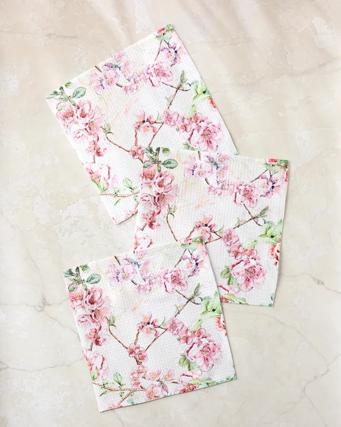 Three folded Pascale Dilger "Primroses" napkins, featuring a pink floral pattern with delicate branches and blossoms, are arranged on a light marble surface, adding style and function to your table.