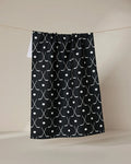 The GEOMETRY Puck tea towel is ultra-absorbent, showcasing a white crossed hockey stick and circle pattern on black, hanging on a string against a neutral background.