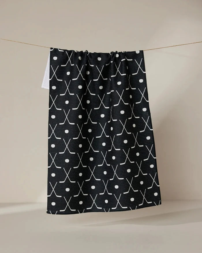 The GEOMETRY Puck tea towel is ultra-absorbent, showcasing a white crossed hockey stick and circle pattern on black, hanging on a string against a neutral background.