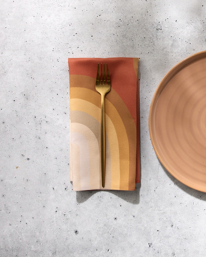 A gold fork rests on a folded GEOMETRY Pumpkin Spice reusable napkin with an earth-toned rainbow pattern, next to a peach ceramic plate on a light gray textured surface.