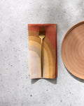 A gold fork rests on a folded GEOMETRY Pumpkin Spice reusable napkin with an earth-toned rainbow pattern, next to a peach ceramic plate on a light gray textured surface.