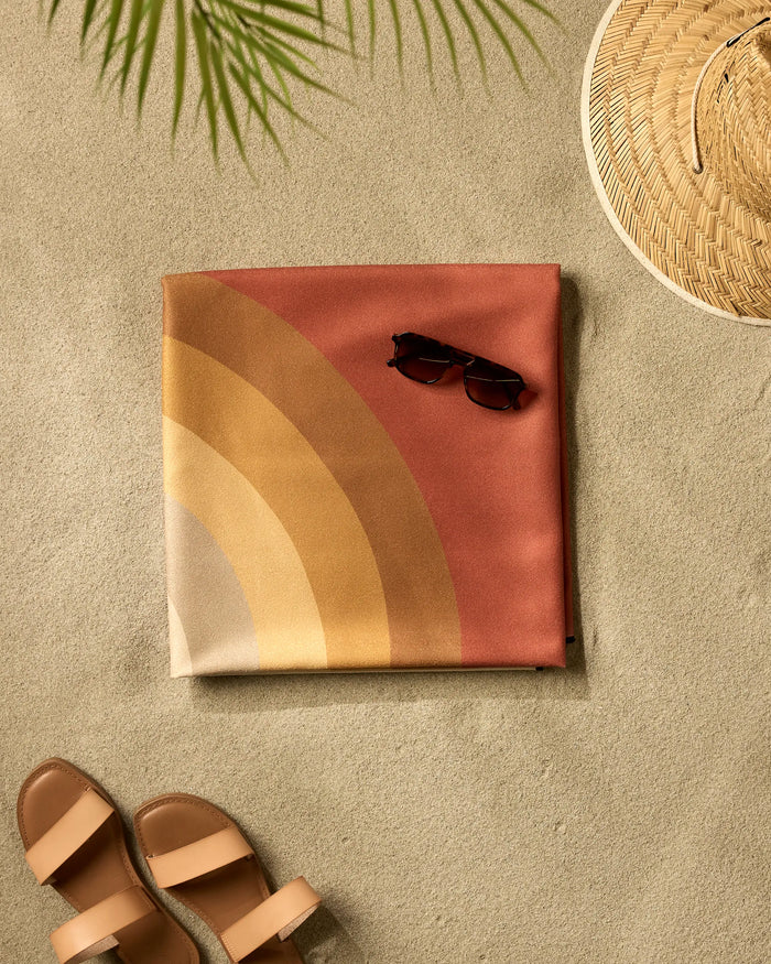 The GEOMETRY "Pumpkin Spice" quick-drying towel, with warm rainbow stripes, is arranged on the sand beside brown sunglasses, a woven straw hat, tan sandals, and topped with a green palm frond.