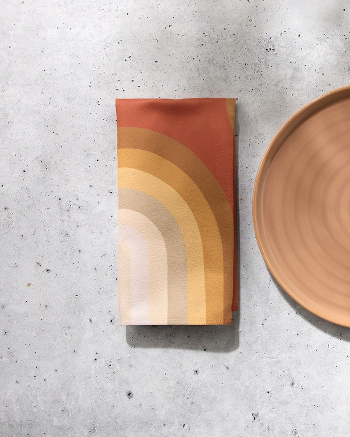 A folded GEOMETRY Pumpkin Spice reusable napkin with a retro rainbow pattern in warm orange, yellow, and cream rests beside a round peach plate on a light gray speckled surface.