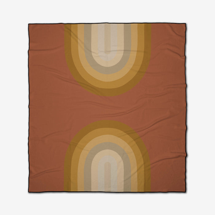 The GEOMETRY Pumpkin Spice recycled beach blanket is square, with a rust background and two mirrored retro rainbow arches in mustard, gold, beige, and tan at the top and bottom. The fabric has subtle wrinkles for a relaxed look.