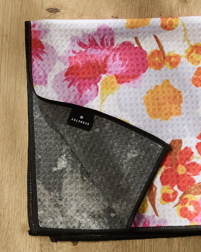 The Geometry Pup Florals Paw Towel, featuring a vibrant floral pattern on one side and solid gray on the back, lies folded on a wooden surface to display both sides. A black label with white text is visible in the corner.