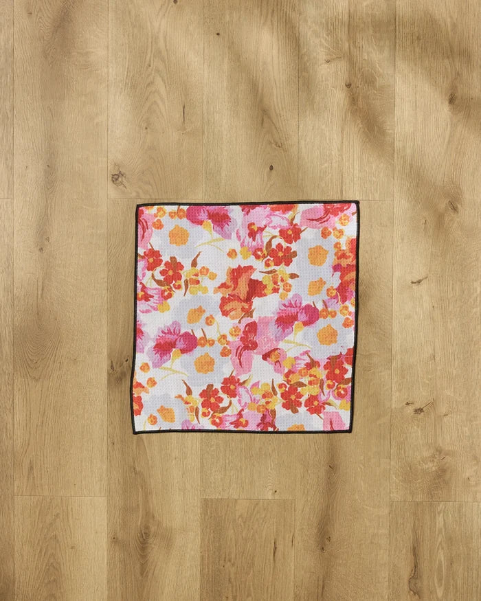The Geometry Pup Florals Paw Towel, featuring a vibrant red, orange, yellow, and pink floral design with a dark border, lies on a light wood floor. This ultra-absorbent towel combines charm and function for pet care.