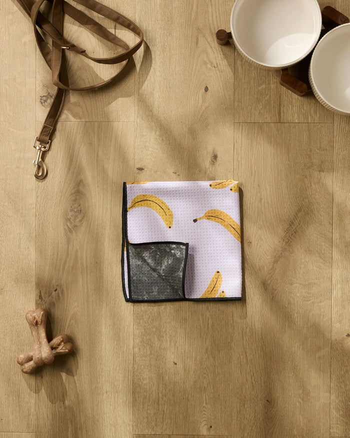 The Geometry Pupnana folded dog bandana rests on a wooden floor beside a leash, two ceramic bowls, an ultra-absorbent dog towel, and a chew bone toy.