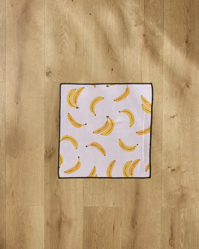 The Geometry Pupnana dog towel features yellow bananas on a white background with a black border. This ultra-absorbent square towel is perfect for drying your pet and looks great flat on any light wooden floor.