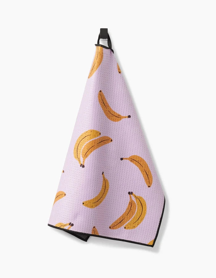 Geometry’s Pupnana kitchen towel features ultra-absorbent Geoweave fabric, a light purple color with a yellow banana pattern, and hangs from a black loop against a white background.
