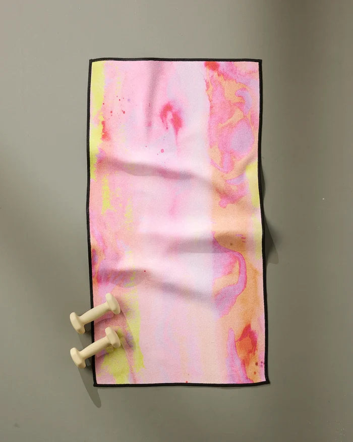 The GEOMETRY Purity towel, ultra-absorbent and fast-drying with pastel pink, yellow, and orange abstract patterns, lies flat on a gray surface. Two small beige dumbbells rest in the bottom left corner.