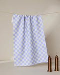 A Purple Picnic recycled kitchen towel by GEOMETRY hangs on a string against a beige background, with two brown mills on a white surface in the lower right corner.