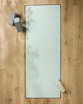 The Rebecca Woolbright Quill Blue yoga mat, featuring a white abstract pattern, is laid on a wooden floor with a non-slip towel, two gray blocks, and a white water bottle.