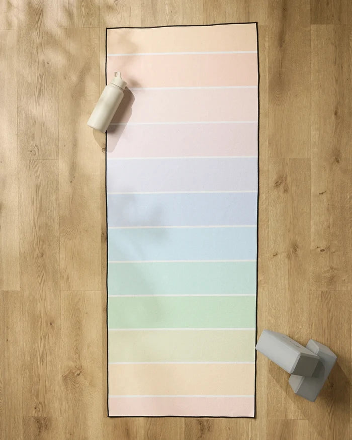 The GEOMETRY Rainbow Pastel yoga mat, featuring horizontal pastel stripes, is spread on a wooden floor with a non-slip towel. A water bottle and two gray yoga blocks sit near the mat’s top left and bottom right corners.