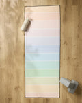 The GEOMETRY Rainbow Pastel yoga mat, featuring horizontal pastel stripes, is spread on a wooden floor with a non-slip towel. A water bottle and two gray yoga blocks sit near the mat’s top left and bottom right corners.